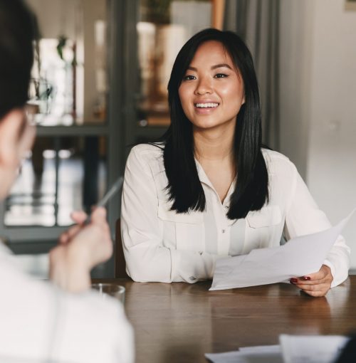 Business, career and placement concept - joyful asian woman smiling and holding resume while sitting in front of directors during corporate meeting or job interview